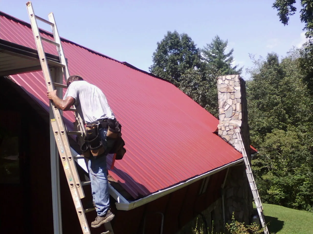 Roofer on a ladder beside a red metal roof and stone chimney — Soffit & Fascia Repair by Roofing Brothers in Nokomis